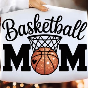 May include: White pillow with the words "Basketball MOM" in black script and block letters. A basketball with a heart and sparkles is inside a net. The pillow is held by a person.
