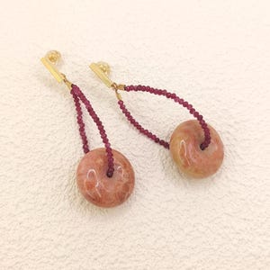 Può includere: Un paio di orecchini pendenti con una pietra circolare rosa-rossastra con un foro al centro. Gli orecchini hanno barre color oro nella parte superiore, collegate alla pietra da un filo di piccole perline rosso scuro. Gli orecchini sono esposti su una superficie bianca.