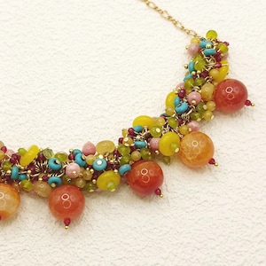 Può includere: Una collana colorata con una catena dorata e un gruppo di piccole perline in vari colori, tra cui giallo, verde, rosa e turchese. Grandi perline rotonde, arancione-rosse, sono sparse nel design, creando un accessorio vibrante e accattivante.