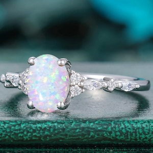 May include: A silver ring featuring an oval, iridescent opal center stone. The ring is accented with smaller, clear gemstones on either side of the opal and along the band. The ring is set against a blurred, teal background.