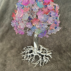 May include: A handcrafted wire tree sculpture with a silver trunk and roots, supporting a canopy of colourful, translucent flowers in shades of pink, purple, blue, and gold. The tree is a decorative item.