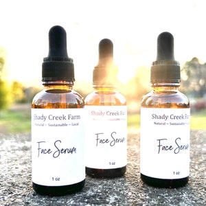 amber glass dropper bottle with white label that reads Face Serum 1 oz. Bottles are sitting outside on concrete table with sun in background shining through bottles with a glowing hue.