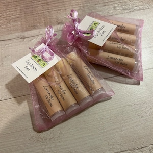 May include: A set of five lavender-scented lip balms in a pink organza bag. The lip balms are packaged in clear tubes with brown labels that read "Lavender Lip Balm". The bag is tied with a pink ribbon.