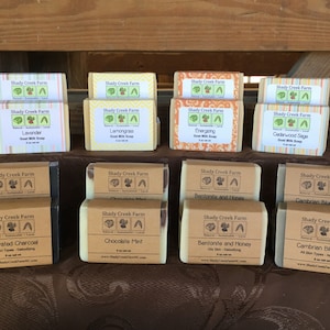 May include: A display of Shady Creek Farm goat milk soaps, each labeled with different scents like Lavender, Lemongrass, and Activated Charcoal. The soaps are wrapped in paper with the brand's logo and product information. The soaps are displayed on a brown surface.