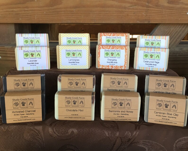 May include: A display of Shady Creek Farm goat milk soaps, each labeled with different scents like Lavender, Lemongrass, and Activated Charcoal. The soaps are wrapped in paper with the brand's logo and product information. The soaps are displayed on a brown surface.