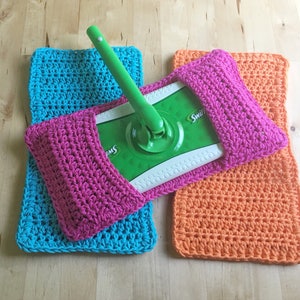 May include: Three crocheted mop head covers in pink, turquoise, and orange. The covers are made with a simple stitch pattern and have a pocket to hold the mop head.