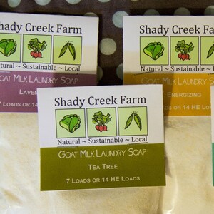 May include: Four different varieties of goat milk laundry soap from Shady Creek Farm. The labels are green, yellow, purple, and white with illustrations of vegetables and the text "Natural ~ Sustainable ~ Local". Each label lists the scent of the soap and states "7 Loads or 14 HE Loads".