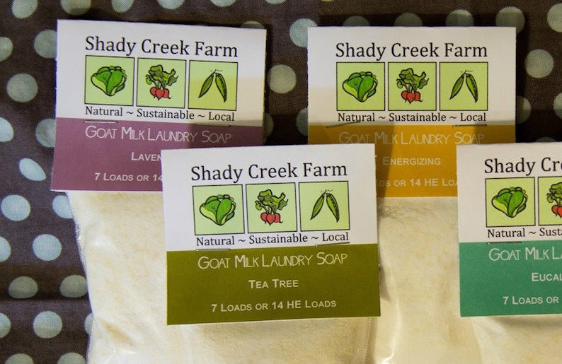 May include: Four different varieties of goat milk laundry soap from Shady Creek Farm. The labels are green, yellow, purple, and white with illustrations of vegetables and the text "Natural ~ Sustainable ~ Local". Each label lists the scent of the soap and states "7 Loads or 14 HE Loads".