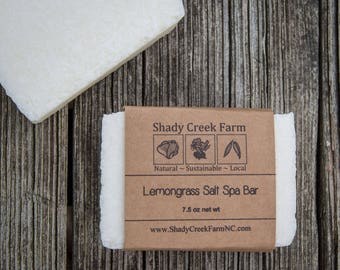May include: A white Lemongrass Salt Spa Bar from Shady Creek Farm, wrapped in a brown paper label. The label includes the text "Natural ~ Sustainable ~ Local" and the bar weighs 7.5 oz.