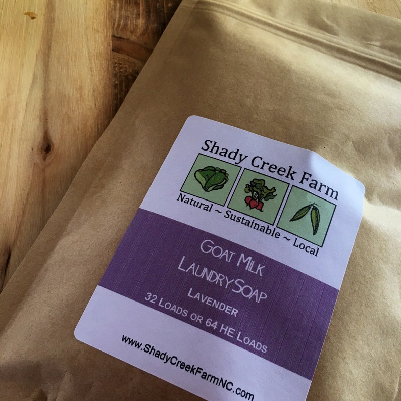May include: A brown paper pouch containing goat milk laundry soap with a lavender scent. The label reads Shady Creek Farm, Natural ~ Sustainable ~ Local, Goat Milk Laundry Soap, Lavender, 32 Loads or 64 HE Loads, and www.ShadyCreekFarmNC.com.