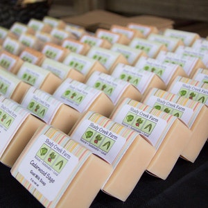 May include: A close-up of a stack of handmade goat milk soap bars with colorful labels. The labels feature the text "Shady Creek Farm" and list the scents "Lavender" and "Cedarwood Sage".