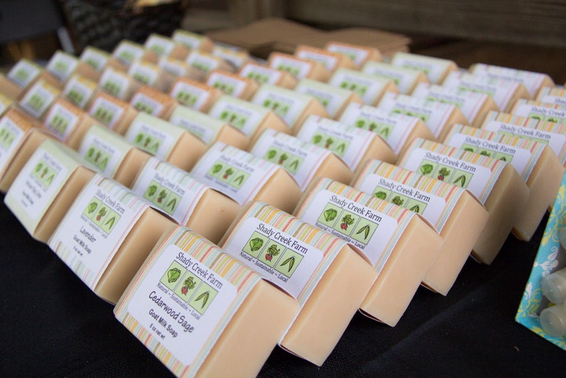 May include: A close-up of a stack of handmade goat milk soap bars with colorful labels. The labels feature the text "Shady Creek Farm" and list the scents "Lavender" and "Cedarwood Sage".
