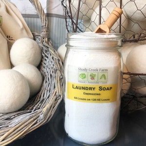 May include: A glass jar filled with white laundry soap powder. The jar has a yellow label that reads "Shady Creek Farm Laundry Soap Energizing 64 Loads or 128 HE Loads". The jar is sitting on a dark blue surface. There are white wool dryer balls in a wicker basket and a wire basket in the background.
