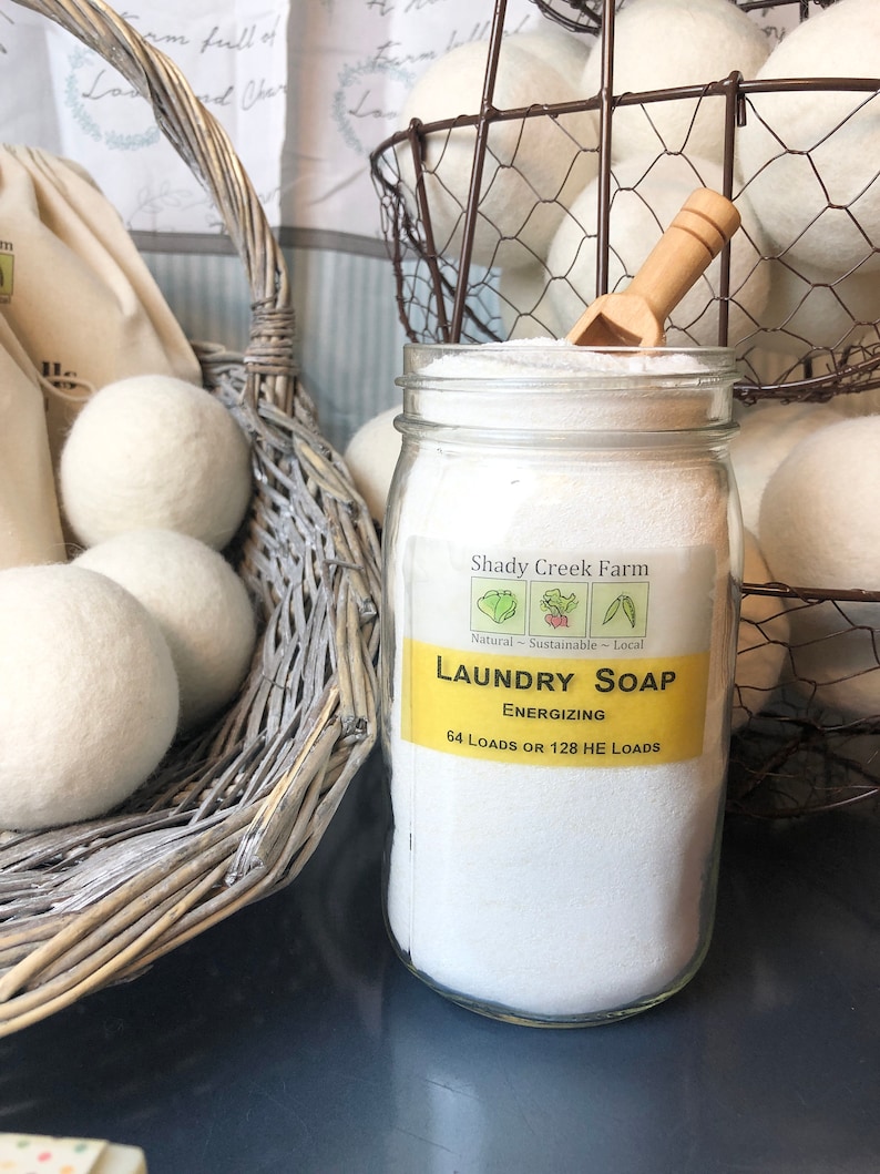 May include: A glass jar filled with white laundry soap powder. The jar has a yellow label that reads "Shady Creek Farm Laundry Soap Energizing 64 Loads or 128 HE Loads". The jar is sitting on a dark blue surface. There are white wool dryer balls in a wicker basket and a wire basket in the background.