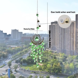 May include: A decorative suncatcher with a sun and moon design, adorned with green crystals. The suncatcher is 14.96 inches long and 2.75 inches wide. It includes a small vial that can hold ashes and hair.