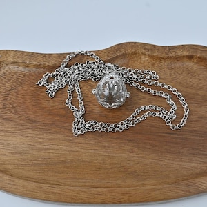 Può includere: Una collana color argento con un pendente a forma di sfera cava. La sfera presenta un intricato design traforato. La catena argentata è ad anello su una superficie di legno.