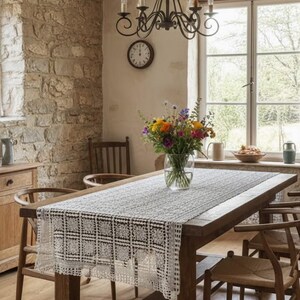 May include: A dining room scene featuring a wooden table with a white lace runner. A vase of colorful flowers sits in the center. The room has a stone wall, wooden chairs, and a wrought iron chandelier. A clock hangs on the wall.