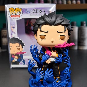 Puede incluir: Una figura de vinilo personalizada de un personaje con cabello negro, vistiendo un chándal negro y naranja, rodeado de numerosas manos azules esculpidas que emergen de una base circular, con un acento rosa cerca del cuello.