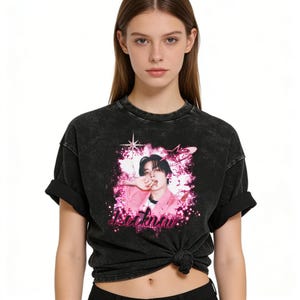 May include: Black acid wash t-shirt featuring a graphic of a person in a pink jacket, with pink accents and the name "Leeknow". The image includes stars and a pink background.