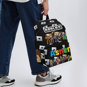 May include: Black backpack featuring the Roblox logo and characters, with the name "Asher" in colorful letters. The backpack has a black background with white square patterns and a front pocket.