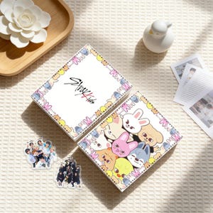 以下が含まれることがあります： 「Stray Kids」の文字が入った白いボックスセット。ボックスは漫画の動物のキャラクターで装飾されています。グループ写真が入った透明なアクリルキーホルダーも2つ見えます。