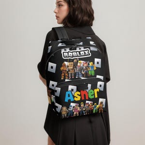 May include: Black backpack featuring the Roblox logo and characters, with the name "Asher" in colorful letters. The backpack has a repeating pattern of white squares and a top handle.