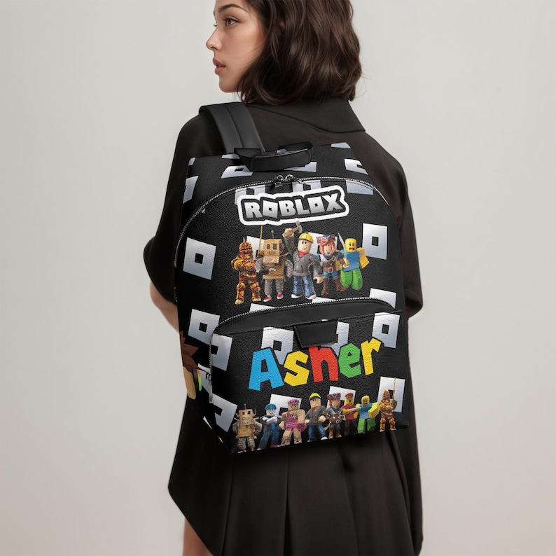 May include: Black backpack featuring the Roblox logo and characters, with the name "Asher" in colorful letters. The backpack has a repeating pattern of white squares and a top handle.