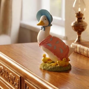 Puede incluir: Figura decorativa de pato con un gorro azul y un chal rosa con lunares. El pato está rodeado de tres patitos amarillos sobre un trozo de hierba verde. La figura está colocada sobre una superficie de madera.