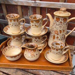 Może przedstawiać: Ceramiczny zestaw do kawy z nakrapianą brązowo-kremową glazurą. Zestaw zawiera dzbanek do kawy, filiżanki, spodki, śmietankę i małą miseczkę, wszystko ułożone na drewnianej tacy.