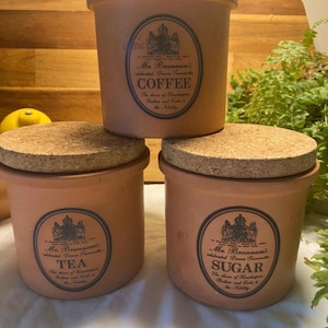 May include: Three terracotta kitchen canisters with cork lids. Each canister has a label with the text "Mr. Brannams" and the words "Coffee", "Tea", and "Sugar". The canisters are stacked on top of each other.