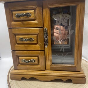 May include: Wooden jewelry box with three drawers and a mirrored door. The box is made of light brown wood and features ornate metal drawer pulls. The door has a floral design etched into the glass. The box sits on a wooden slice.