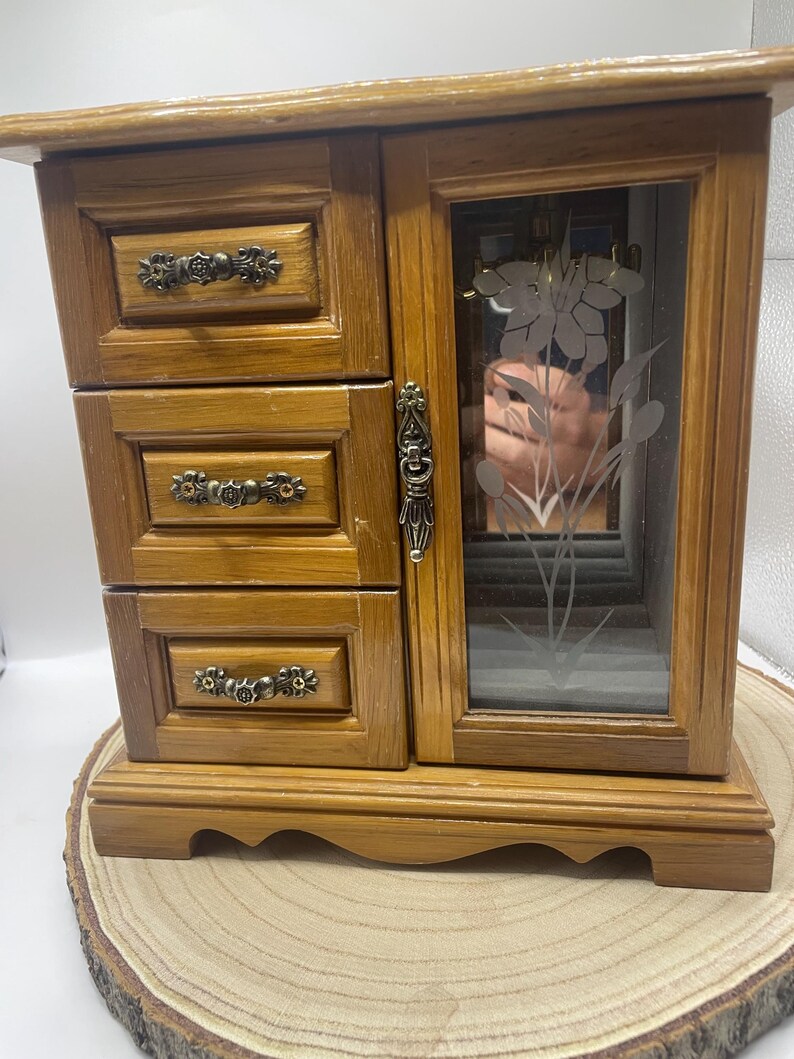 May include: Wooden jewelry box with three drawers and a mirrored door. The box is made of light brown wood and features ornate metal drawer pulls. The door has a floral design etched into the glass. The box sits on a wooden slice.