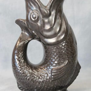 Puede incluir: Jarra de cerámica marrón oscuro con forma de pez. La jarra tiene una boca ancha y una cola que forma un asa. El pez tiene escamas y aletas, y se apoya sobre una pequeña base redonda. Aproximadamente 20 cm de altura.