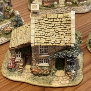 Puede incluir: Figurita de una casa en miniatura con fachada de piedra, tejado de tejas amarillas y una pequeña chimenea. La casa tiene una puerta verde, ventanas pequeñas y está rodeada de vegetación. La base es de color verde claro.