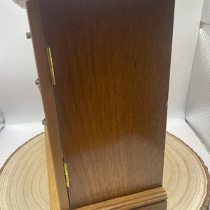 May include: A small, wooden jewelry box with a hinged door and brass-colored hardware. The box is a warm, medium brown color and sits on a wooden base. The box is displayed on a wooden slice.