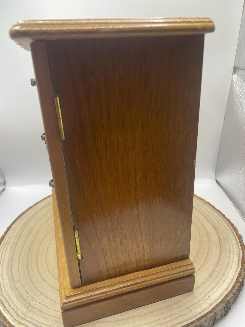 May include: A small, wooden jewelry box with a hinged door and brass-colored hardware. The box is a warm, medium brown color and sits on a wooden base. The box is displayed on a wooden slice.