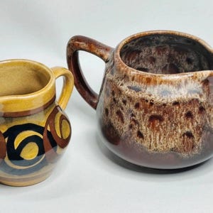Könnte beinhalten: Zwei Keramikkrüge mit Henkeln und Ausgüssen. Der kleinere Krug hat eine gelbe Basis mit braunen und schwarzen kreisförmigen Mustern. Der größere Krug hat eine glänzende braune Glasur mit einer texturierten, gesprenkelten cremefarbenen und braunen Oberfläche.