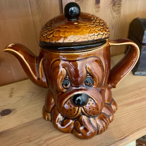 Pode incluir: Uma chaleira de cerâmica marrom em forma de cabeça de cão, com uma tampa com design de bolota texturizada e um botão preto. A chaleira tem um acabamento brilhante e está em uma prateleira de madeira.