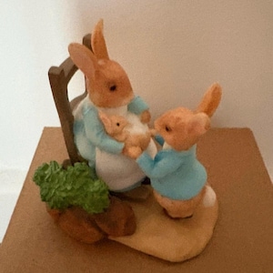 May include: A figurine depicting a family of rabbits. One rabbit sits in a chair, holding a baby rabbit. Another rabbit stands nearby. The rabbits wear blue jackets and are set on a tan base with a green vegetable.