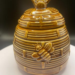 May include: A ceramic honey pot with a lid, featuring a bee-themed design. The pot is brown with horizontal lines and a raised bee emblem on the front. The lid is topped with a bee figurine. The pot has a round shape and is likely used for storing honey.
