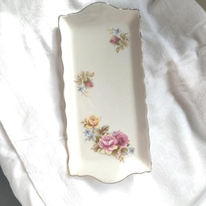 Puede incluir: Una bandeja rectangular de cerámica blanquecina con borde festoneado y ribete dorado. Está decorada con racimos de rosas rosas, amarillas y azules. Una pieza decorativa para servir o exhibir, de unos 20 cm de largo.
