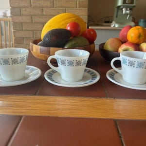 May include: Three white teacups and saucers with a blue floral pattern. The cups and saucers are arranged on a brown tiled surface. A wooden bowl of fruit is in the background.