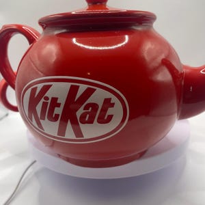 May include: A red ceramic teapot featuring the Kit Kat logo in white. The teapot has a rounded body, a handle, a spout, and a lid with a small knob. The teapot is on a white surface.