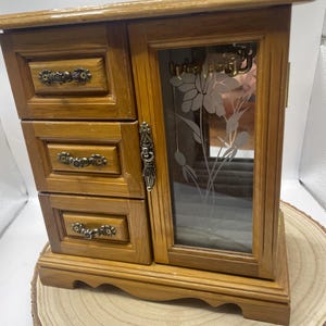 May include: Wooden jewelry box with three small drawers on the left and a glass door on the right. The glass door has a floral design etched into it. The box is made of light brown wood and sits on a wooden base.