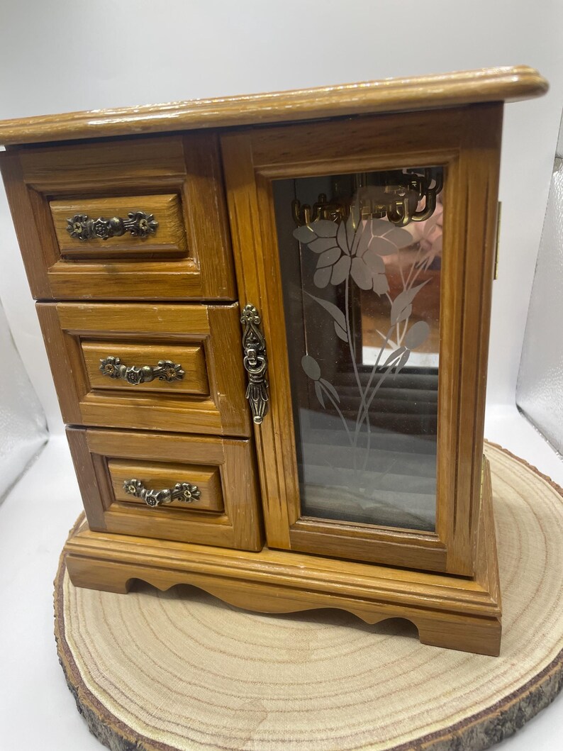 May include: Wooden jewelry box with three small drawers on the left and a glass door on the right. The glass door has a floral design etched into it. The box is made of light brown wood and sits on a wooden base.