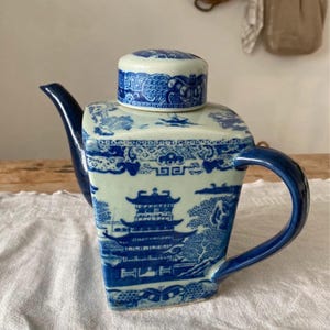 May include: A blue and white porcelain teapot with a square body and a curved handle. The teapot features a detailed landscape design with pagodas and trees. The lid is also decorated with a blue and white pattern.