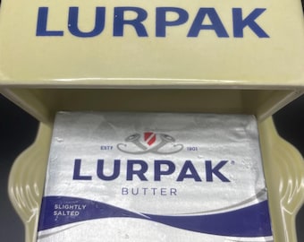 May include: A light yellow butter dish with the word "LURPAK" in blue lettering on the lid. Inside the dish is a silver and blue wrapped block of butter, also labeled "LURPAK". The butter is slightly salted.