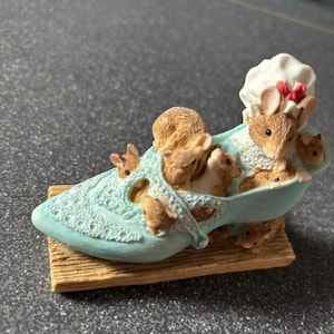 May include: A charming figurine depicting a shoe-shaped bathtub occupied by several brown mice. The blue bathtub rests on a wooden base. One mouse wears a white chef's hat with a red bow. A delightful decorative item.