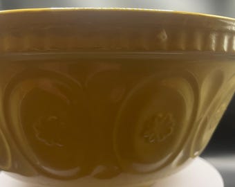 Puede incluir: Un cuenco amarillo para mezclar con patrones florales y circulares en relieve. El cuenco tiene un borde ancho y una forma redondeada. El cuenco está sobre una superficie blanca.
