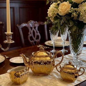 Puede incluir: Un juego de té dorado con detalles florales, que incluye una tetera, una cremera y un azucarero, se encuentra sobre un mantel blanco. Un ramo de rosas blancas y una vela encendida en un candelabro plateado completan la elegante escena.